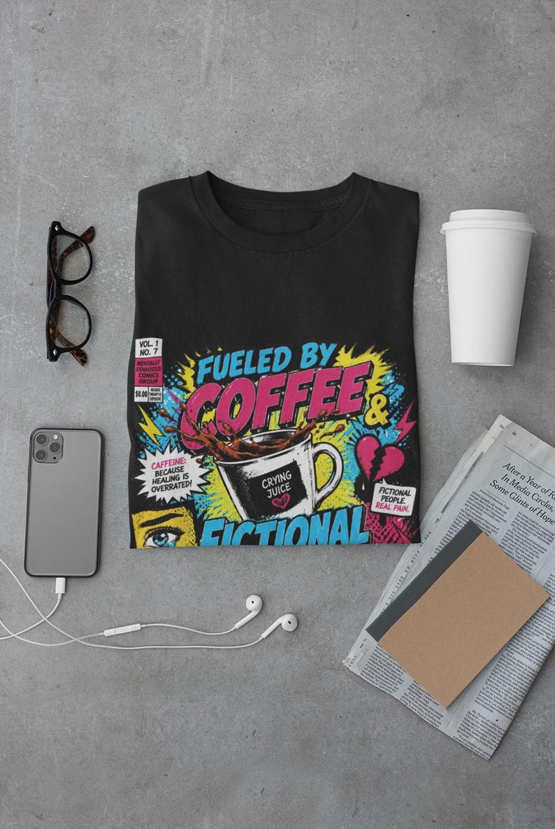 Aesthetic flat lay of black Fueled by Coffee and Fictional Trauma bookish tee with glasses, notebook, phone and earphones, bookish gift idea styling