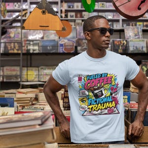 Man at outdoor book market wearing white Fueled by Coffee and Fictional Trauma bookish graphic tee, surrounded by stacked books, book lover lifestyle