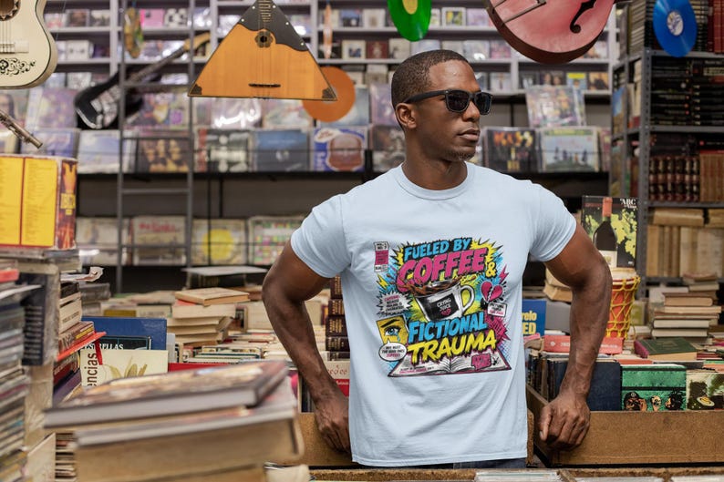 Man at outdoor book market wearing white Fueled by Coffee and Fictional Trauma bookish graphic tee, surrounded by stacked books, book lover lifestyle
