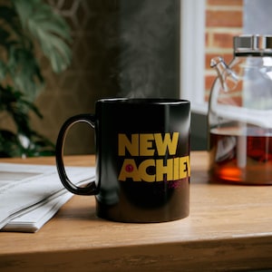 May include: A black ceramic mug with the words "NEW ACHIE" in gold lettering. The mug is on a wooden surface, with steam rising from it. A glass teapot with tea is in the background.