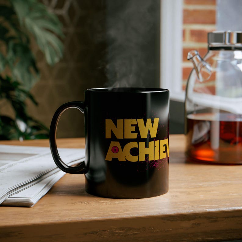 May include: A black ceramic mug with the words "NEW ACHIE" in gold lettering. The mug is on a wooden surface, with steam rising from it. A glass teapot with tea is in the background.