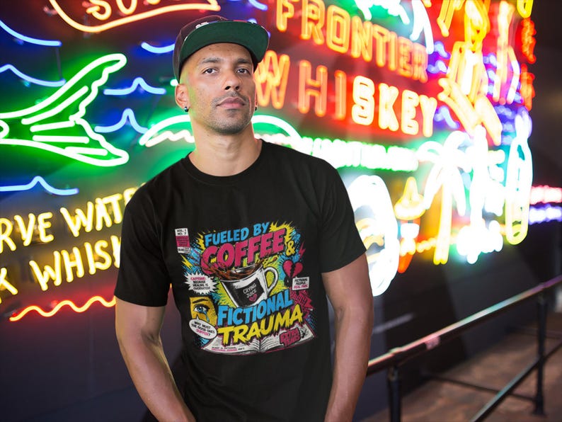Man wearing black Fueled by Coffee and Fictional Trauma bookish tee in front of colorful neon bar signs, vibrant nightlife lifestyle photo