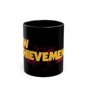 Clean, professional product photograph of a black ceramic mug featuring "NEW ACHIEVEMENT" text in bright gold lettering against a pure white background. The straight-on angle clearly displays the full text and demonstrates the high contrast between the glossy black ceramic and metallic gold lettering. This minimalist presentation highlights the product details without distractions, allowing shoppers to see the exact design, proportions, and finish quality.