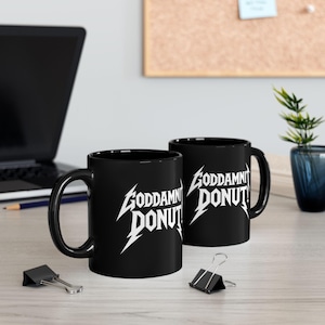 Black ceramic mug with bold white text in a Metallica-style font that reads “Goddamnit Donut,” inspired by Dungeon Crawler Carl. The mug is set on a modern kitchen counter, perfect for fans of LitRPG, Princess Donut, and Matt Dinniman's cult book series. Ideal for coffee or tea lovers who enjoy a mix of humor, chaos, and fandom in their daily routine.