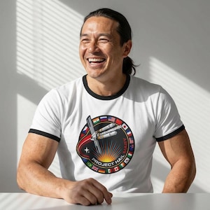 Puede incluir: Camiseta blanca con cuello y mangas negras. La camiseta presenta un diseño gráfico circular con un diseño de transbordador espacial, el texto "PROJECT HAIL MARY" y banderas de varios países.