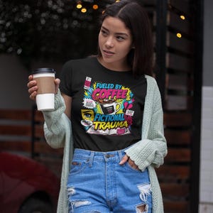 Woman holding coffee cup wearing black Fueled by Coffee and Fictional Trauma bookish tee with denim shorts, string lights background, bookish gift shirt