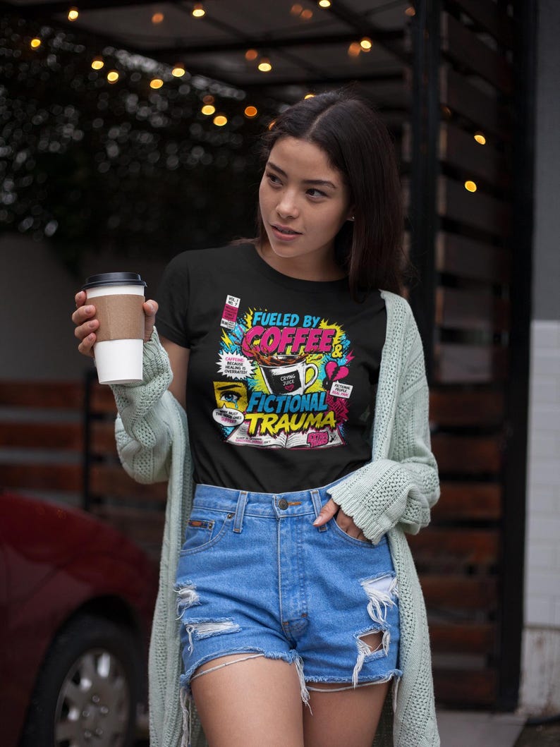 Woman holding coffee cup wearing black Fueled by Coffee and Fictional Trauma bookish tee with denim shorts, string lights background, bookish gift shirt