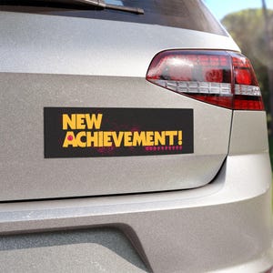 Peut inclure: Un autocollant de pare-chocs noir et jaune avec le texte "NEW ACHIEVEMENT!" dans une police audacieuse et stylisée. L'autocollant a un effet de projection rose et blanc.