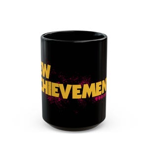 May include: Black ceramic mug with the words "NEW ACHIEVEMENT" in gold lettering. The text is overlaid on a pink splatter design. The mug is cylindrical and has a glossy finish.