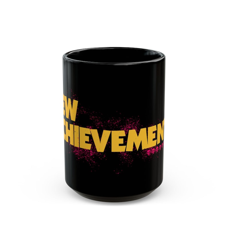May include: Black ceramic mug with the words "NEW ACHIEVEMENT" in gold lettering. The text is overlaid on a pink splatter design. The mug is cylindrical and has a glossy finish.