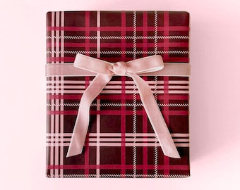 May include: A rectangular gift box wrapped in burgundy plaid paper with a light pink ribbon and bow. The box is centered on a light pink background, creating a festive and elegant presentation.