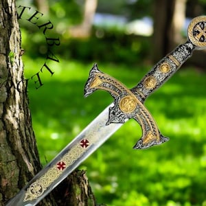 May include: A silver and gold sword with a cross-shaped pommel and intricate details. The sword is resting on a green grassy surface.