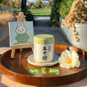 May include: A wooden tray holds a small painting of a cartoon bunny, a jar with Japanese text, and a white flower. The jar is white with a green lid and a label with Japanese characters. The text "40g & 100g" is written in green.