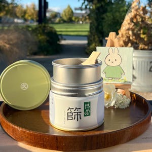 May include: A metal container with a lid and a wooden scoop, likely for tea or a similar product. The container has Japanese text on the label. A small painting of a bunny is in the background.