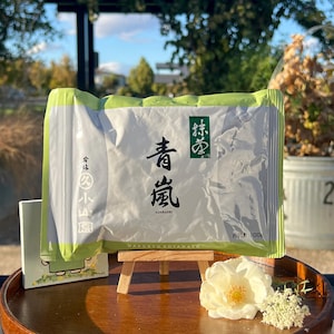 May include: A package of Japanese matcha tea with green and white packaging. The package has Japanese characters and the word "ADARASHI" printed on it. The package is displayed on a small wooden easel with a white flower and small white flowers.