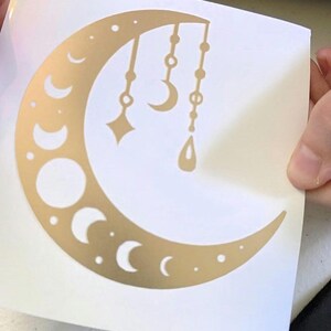 Puede incluir: Pegatina dorada con forma de media luna con varias fases lunares y colgantes. El dise&ntilde;o incluye una estrella, una media luna y una forma de l&aacute;grima, suspendidas de la parte superior de la luna.