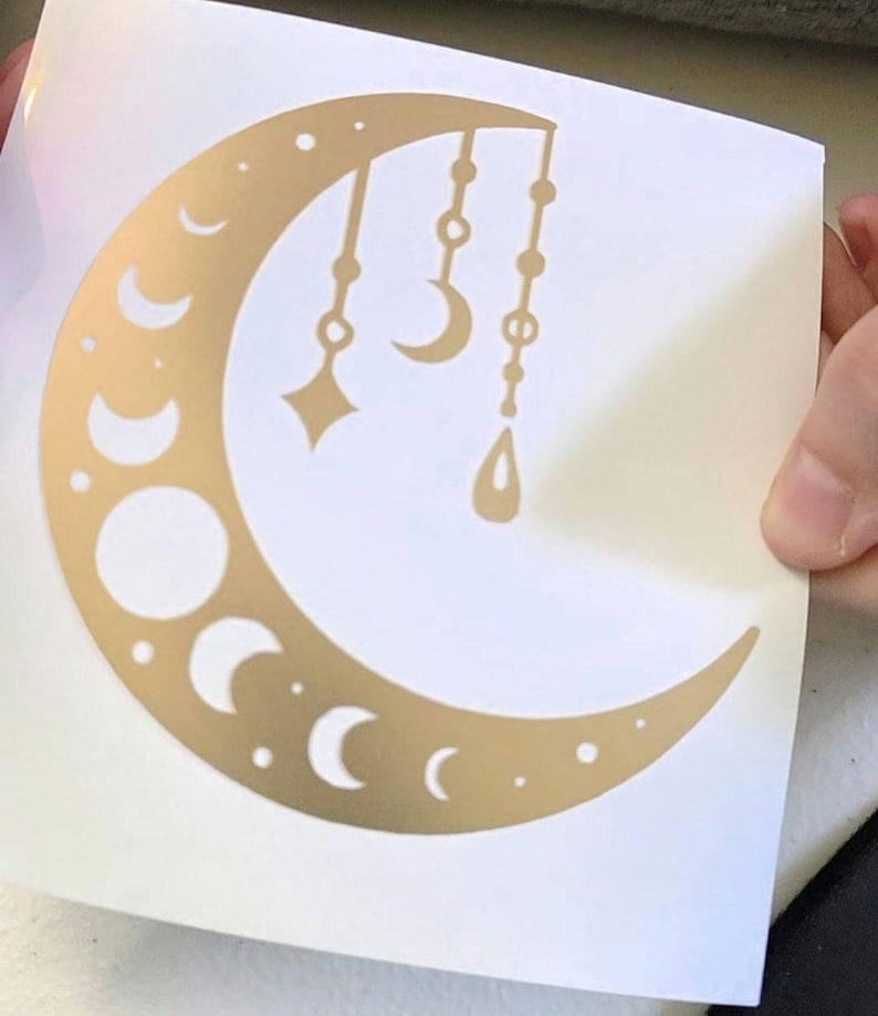 Puede incluir: Pegatina dorada con forma de media luna con varias fases lunares y colgantes. El dise&ntilde;o incluye una estrella, una media luna y una forma de l&aacute;grima, suspendidas de la parte superior de la luna.