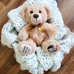 May include: A light blue and white crocheted baby blanket with a brown teddy bear wearing a gold ribbon. The blanket has a personalized tag with the text "Theodore Waylon 2021".