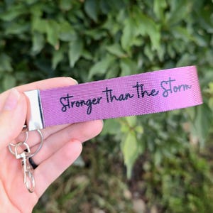 May include: A purple key fob with the text "Stronger than the Storm" in black script. The key fob has a silver metal clasp and key ring. The background is blurred green foliage.