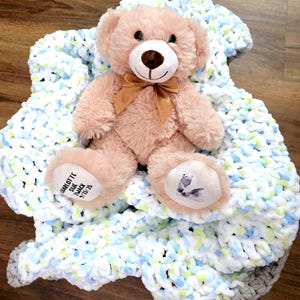 May include: A plush, light brown teddy bear with a brown bow tie sits atop a soft, handmade blanket. The blanket is white, blue, and green. The bear's foot has text that reads "CHARLOTTE SUE MACK 9/23/25".