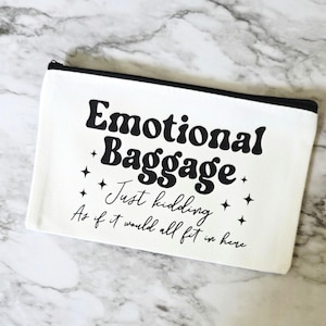 May include: White canvas zippered pouch with the text "Emotional Baggage" in a bold, retro font. Below the text are the words "Just kidding" and "As if it would all fit in here" in a smaller, cursive font. Black zipper.