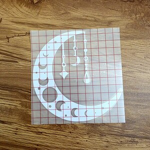 Puede incluir: Calcoman&iacute;a de vinilo blanca con forma de media luna con varias fases lunares y elementos decorativos colgantes. El dise&ntilde;o incluye c&iacute;rculos, medias lunas y formas de l&aacute;grima, creando un tema celestial. La calcoman&iacute;a est&aacute; sobre un respaldo de cuadr&iacute;cula transparente.