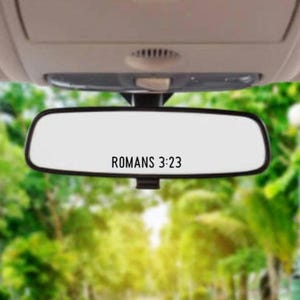 May include: A rearview mirror with the text "ROMANS 3:23" printed on it. The mirror is hanging from the ceiling of a car. The background is a blurry image of green trees.
