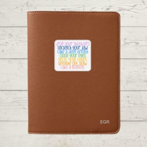 May include: A brown leather journal with a sticker on the front that says "drop your shoulders, unclench your jaw, take a deep breath, close your eyes, relax your hands, breathe out slow, take a moment". The journal has the letters "EGR" embossed on the bottom right corner.