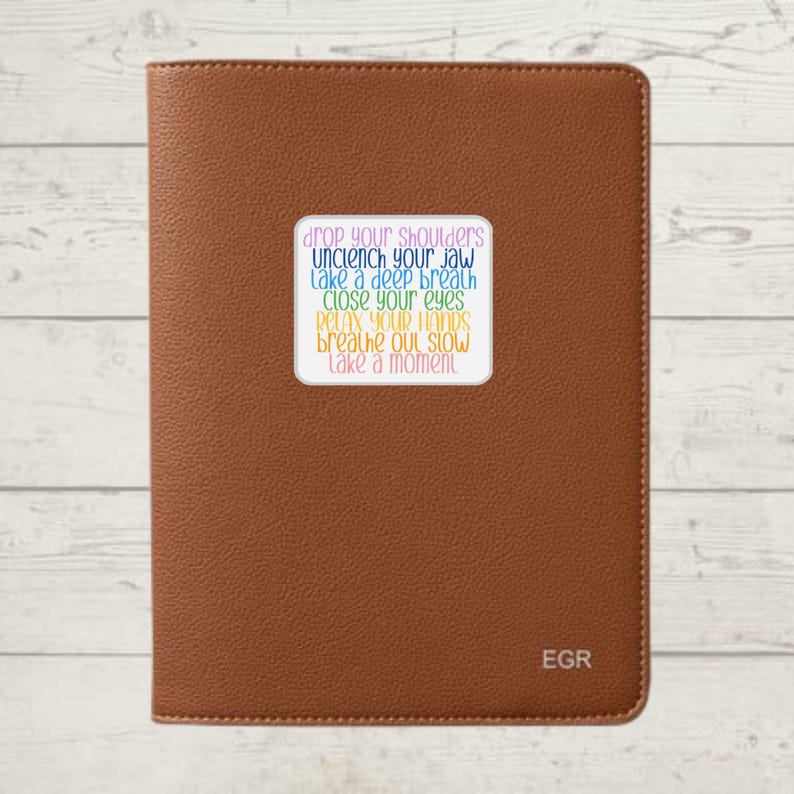 May include: A brown leather journal with a sticker on the front that says "drop your shoulders, unclench your jaw, take a deep breath, close your eyes, relax your hands, breathe out slow, take a moment". The journal has the letters "EGR" embossed on the bottom right corner.