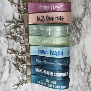 May include: A collection of keychains with fabric straps in various colors, each featuring inspirational phrases in white lettering. Phrases include "Pray First," "Faith Over Fear," and "Heaven Bound." The keychains have silver-colored metal hardware.