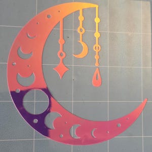 Puede incluir: Un dise&ntilde;o de media luna hologr&aacute;fica con formas celestiales colgantes en tonos naranja, rosa y morado. La luna presenta formas de media luna recortadas y un gran recorte circular. Las formas colgantes incluyen una estrella, una media luna y una l&aacute;grima.
