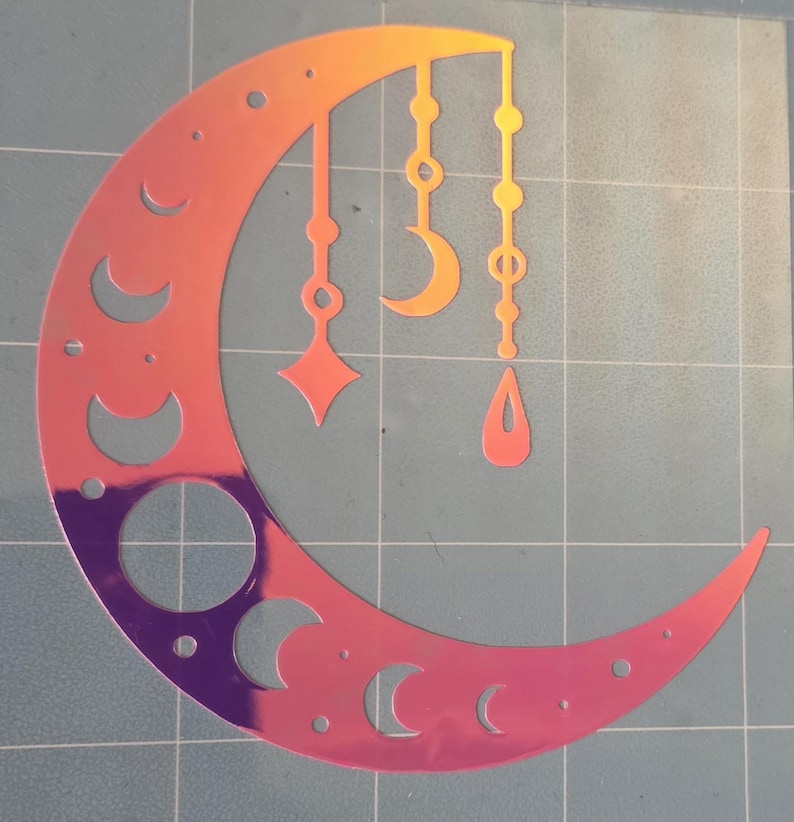 Puede incluir: Un dise&ntilde;o de media luna hologr&aacute;fica con formas celestiales colgantes en tonos naranja, rosa y morado. La luna presenta formas de media luna recortadas y un gran recorte circular. Las formas colgantes incluyen una estrella, una media luna y una l&aacute;grima.