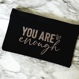May include: Black zippered pouch with the words "YOU ARE enough" in glittery silver lettering. The pouch is rectangular and rests on a marble-patterned surface. The text includes a small heart symbol.
