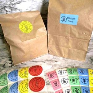 May include: Two brown paper bags with "Gluten Free" labels, one with a blue rectangle label and the other with a yellow circle label. Several sheets of colorful "Gluten Free" stickers are in the foreground, including red, yellow, blue, green, and pink.
