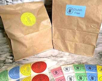 May include: Two brown paper bags with "Gluten Free" labels, one with a blue rectangle label and the other with a yellow circle label. Several sheets of colorful "Gluten Free" stickers are in the foreground, including red, yellow, blue, green, and pink.