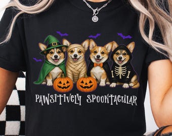 May include: A black t-shirt featuring a graphic of four Corgi dogs dressed in Halloween costumes, including a witch, a mummy, a vampire, and a skeleton. Two orange jack-o'-lanterns sit in front of the dogs, with the text Pawsitively Spooktacular.