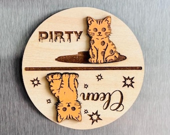 May include: A wooden, circular magnet with a cat theme. The magnet has the words "Dirty" and "Clean" with a cat illustration for each. The "Dirty" cat is upright, and the "Clean" cat is upside down. The magnet is light brown.