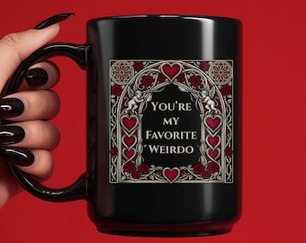 May include: Black ceramic mug with a large handle. The mug features a decorative design with red hearts, roses, and the text "You're My Favorite Weirdo" in a gothic font. The mug is held against a red background.