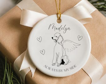 May include: A white ceramic ornament with a gold string, featuring a black line drawing of a poodle with angel wings. The ornament has the name "Madelyn" and the phrase "ALWAYS BY MY SIDE". The ornament is on a wrapped gift.