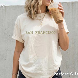 Puede incluir: Camiseta de color marfil con el texto "SAN FRANCISCO" en una fuente verde claro. La camiseta es de corte holgado y tiene las mangas enrolladas. La modelo sostiene un vaso de plástico transparente con una bebida marrón.