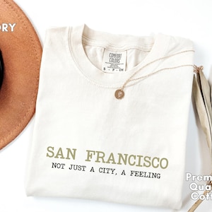 Puede incluir: Camiseta de color marfil con el texto "SAN FRANCISCO NOT JUST A CITY, A FEELING" en verde oliva y negro. La camiseta está hecha de algodón de primera calidad y tiene cuello redondo. Un collar dorado con un colgante descansa sobre la camiseta.
