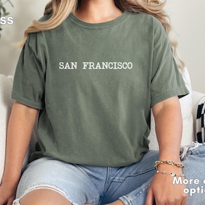 May include: Moss green t-shirt with the text "SAN FRANCISCO" in white. The shirt is a relaxed fit and is paired with blue denim shorts. The image also shows a gold bracelet and a white pillow.