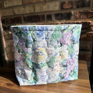 May include: A quilted fabric basket with a floral pattern in shades of blue, purple, green, and yellow. The basket is rectangular and appears to be handmade. The background is a brick wall and a wooden surface.