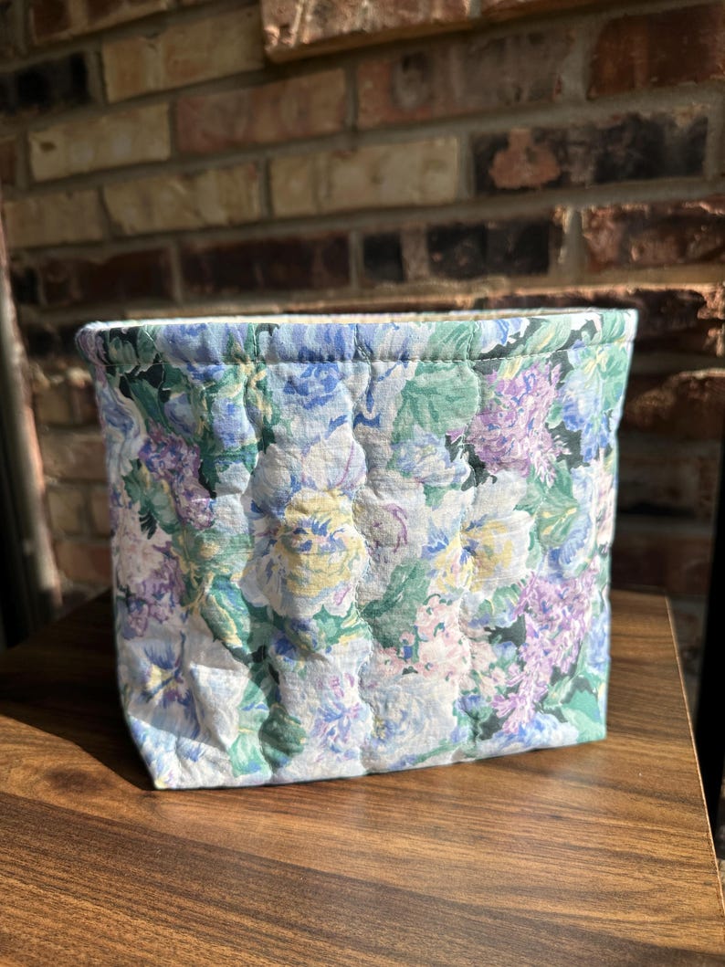 May include: A quilted fabric basket with a floral pattern in shades of blue, purple, green, and yellow. The basket is rectangular and appears to be handmade. The background is a brick wall and a wooden surface.
