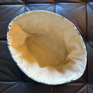 May include: An open, floral-patterned fabric basket with a light beige interior. The exterior features a design of purple, green, and blue flowers. The basket is likely handmade and suitable for storage or decorative use.