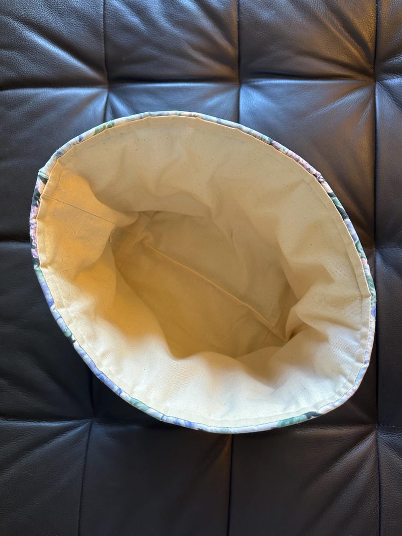 May include: An open, floral-patterned fabric basket with a light beige interior. The exterior features a design of purple, green, and blue flowers. The basket is likely handmade and suitable for storage or decorative use.