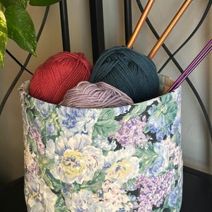 May include: A fabric storage basket filled with yarn and knitting needles. The basket has a floral pattern with white, blue, and purple flowers. Inside are balls of red, teal, and lavender yarn, along with two copper and two purple knitting needles.