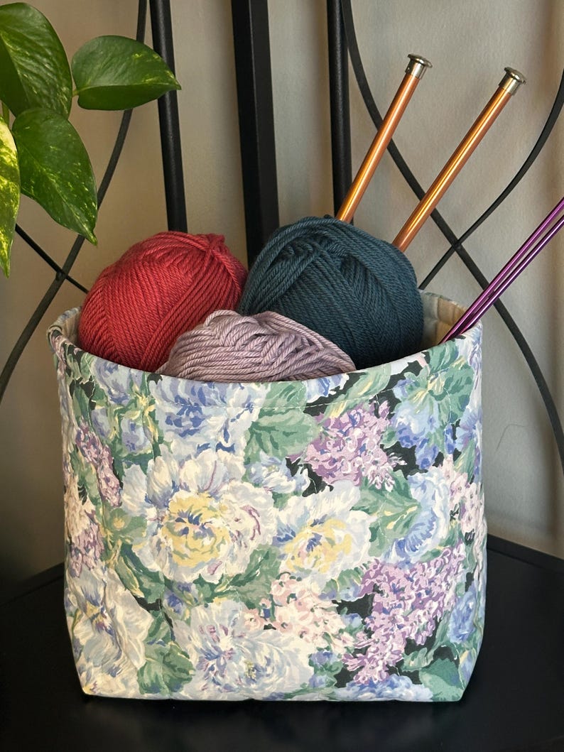 May include: A fabric storage basket filled with yarn and knitting needles. The basket has a floral pattern with white, blue, and purple flowers. Inside are balls of red, teal, and lavender yarn, along with two copper and two purple knitting needles.