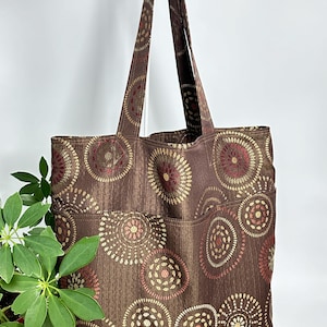 Puede incluir: Una bolsa de mano marrón con un patrón circular en dorado y rojo. La bolsa tiene dos bolsillos en la parte delantera.