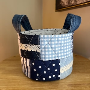 May include: A patchwork fabric basket with denim handles. The basket features a variety of blue fabrics, including denim, polka dots, gingham, and striped patterns. The interior is white with small black polka dots. The basket is sitting on a wooden surface.